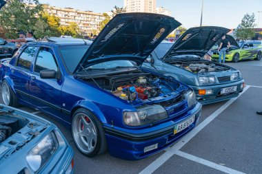 Açık havada kaputu açık mavi hatchback araba, yakınlarda Grayblack motor parçaları sergiliyor. Bir toplantıda araçlara odaklanan uzaktaki insanlar veya Kyiv, Ukrayna - 21 Eylül 2025