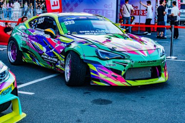 Canlı, kompakt bir yarış arabasının sergilendiği kapalı bir sergi. Pembe ve maviye boyanmış yeşil aksanlı, dinamik soyut tasarımlı, 21 Eylül 2025 'te Ukrayna' nın başkenti Kyiv 'de konumlanmış.