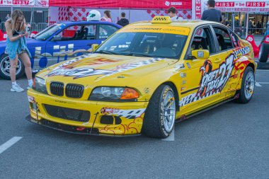 Alevli sarı bir BMW yarış arabası, asfalt bir yüzeye yerleştirilmiş, çeşitli araçlarla çevrili çıkartmalar ve sponsor logolarıyla detaylandırılmış gizli bir tarzda Kyiv, Ukrayna - 21 Eylül 2025