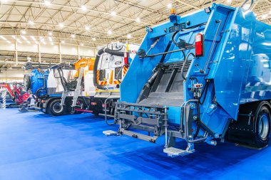 Kapalı alanda endüstriyel ortam, hidrolik kollu ve büyük çöp kutusuyla öne çıkan mavi çöp kamyonu, sarı gri atık toplama aracı, arka planda çeşitli renkli kamyonlar Kyiv, Ukrayna - 14 Ekim 2025