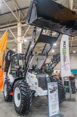 Genişletilmiş yükleyicili modern bir beyaz ve siyah traktörün sergilendiği kapalı sanayi sergisi, bir depoda konumlandırılmış arka plan özellikleri Kyiv, Ukrayna - 14 Ekim 2025