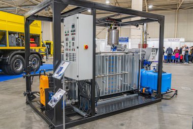 Arka planda karmaşık borular, hortumlar, kontrol paneli ve elektrikli kamyonla donatılmış bir depo ortamında mavi ve gri endüstriyel makineler 14 Ekim 2025