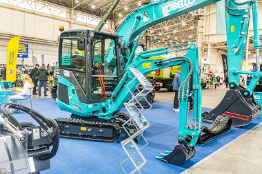 İki KOBELCO ağır makine traktörünün sergilendiği kapalı sergi, mavi halılı bir zeminde kazıcı Ziyaretçiler veya arka planda bulunan eşit ışıklandırılmış uzay zekası Kyiv, Ukrayna - 14 Ekim 2025