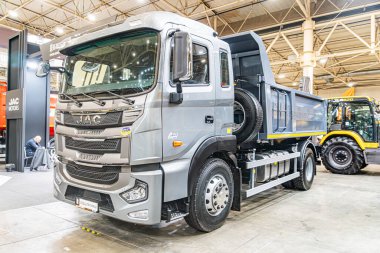 Büyük bir depo galerisinde endüstriyel sergi. Gri damperli kamyon ve sarı inşaat aracı sergileniyor. Parlak, kapalı alanda bile ışıklandırma yapılıyor.