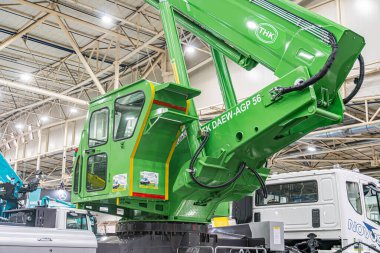 Güvenlik şeritli eklemli yeşil kazıcı arka planda park halindeki kamyon vinci ve 14 Ekim 2025 'te Ukrayna' nın başkenti Kyiv 'in yapay ışıklandırmasının yer aldığı kapalı sanayi bölgesi.