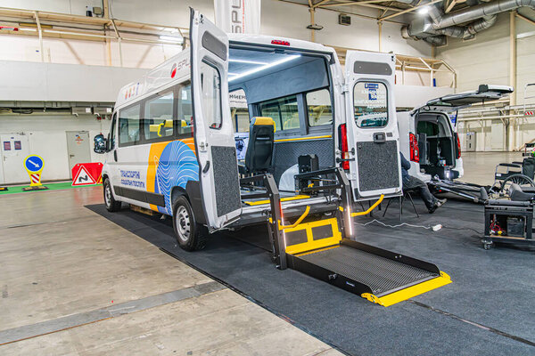 Два автобуса для перевозки людей с ограниченными возможностями, один синий и другой желтый, припаркованы в помещении в промышленной зоне. Оба имеют задние пандусы.