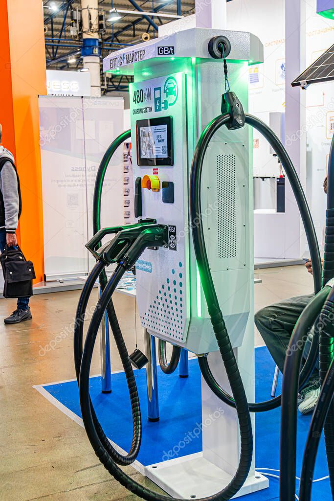 Indoor trade show scene with a white, powered electric charging station featuring green accents and multiple black cables, partially obscuring a person walking away Kyiv, Ukraine - October 14, 2025