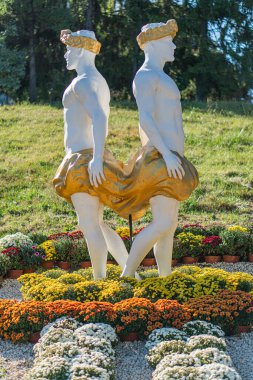 Erkek figürlerinin altın, senkronize, yürüyen heykelleri renkli bir çiçek arkaplanına karşı kaslı gövdeler turuncu ve beyaz çiçekleri, tomurcuklanan bazı Brig Kyiv, Ukrayna - 20 Eylül 2025