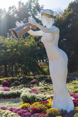 Beyaz deniz kızı Zodiac Aquarius 'un elinde trompet bulunan heykeli, canlı bir bahçede yemyeşil, pembe ve mor çiçekler öğleden sonra veya erken saatlerde Eveni Kyiv, Ukrayna - 20 Eylül 2025