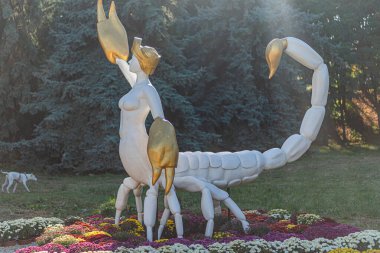 Altın Zodiac Akrep heykeli, canlı mor çiçekleri ve koyu yeşil yaprakları olan bir bahçenin ortasında insansı figür bir ıstakoz pençesi tutuyor, Ukrayna, Kyiv 'e karşı, 20 Eylül 2025