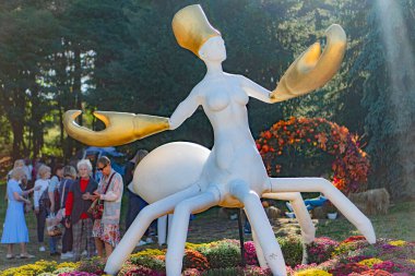 Yeşillik bir bahçede ıstakoz benzeri uzantıları olan büyük, beyaz bir heykel serpiştirilmiş seyirciler parçaya doğal gün ışığı altında hayran kalıyorlar.