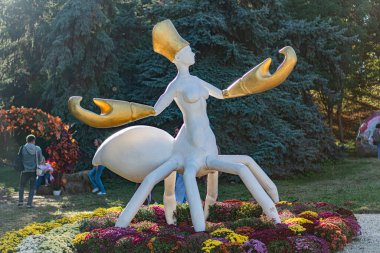 Beyaz plastik zodyak kanseri heykeli, bir ıstakozun tepesinde bir kraliyet figürü yer alıyor, yeşillik içinde göze çarpan ıstakoz detaylandırılmış, mor çiçeklere dayanıyor insanlar Kyiv, Ukrayna - 20 Eylül 2025