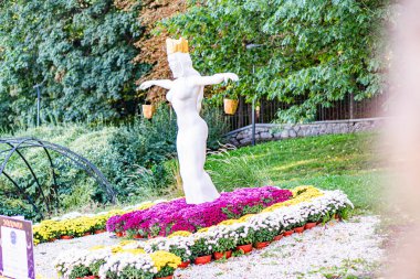 Canlı bir bahçe ortamında beyaz, geometrik, soyut insan figürü heykel, yemyeşil çiçekler arasında, kırmızı, pembe, beyaz, turuncu, ağaçlar tamamen çiçek açıyor, yeşil, siz Kyiv, Ukrayna - 20 Eylül 2025