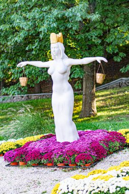 Zodiac Başak Heykeli, yemyeşil çitler, açık bej taş heykel, açık kollar, taç, canlı çiçek yatakları, pembe ve mor çiçek Kyiv, Ukrayna - 20 Eylül 2025