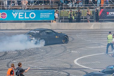 Kyiv Araba Sürükleme Festivali - Dinamik siyah BMW asfalt yolda sürükleniyor, izleyiciler bariyerlerden izliyor, daha fazla araba sıraya diziliyor veya park ediliyor, duman yükseliyor. Kyiv, Ukrayna 06-15-2025