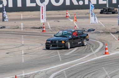 Kyiv Araba Sürükleme Festivali 'nde çok katmanlı toprak yolda sürüklenen siyah BMW, açık gökyüzüne karşı güvenlik konileri, dinamik hareketler, canlı renkler, gerçekçi stil Kyiv, Ukrayna 06-15-2025