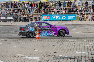 Sürüklenen bir festivalde mor BMW arabası keskin bir sola dönüş yapıyor, seyirciler pist boyunca dizildi, açık mavi gökyüzü Kyiv, Ukrayna 06-15-2025