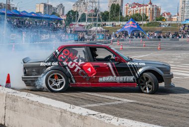 Kırmızı ve siyah BMW, Kyiv Araba Sürükleme Festivali sırasında bir yarış pistinde sürükleniyor. Seyirciler katmanlardan izliyor. Lastiklerin arkasından duman görünüyor.