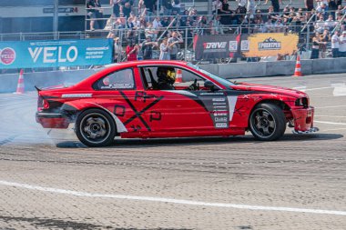 Kırmızı BMW arabası bir yarış pistinde sürüklenirken sarı takım elbiseli sürücü virajın etrafında manevra yapıyor, arka lastikler patlatılıyor diğer arabalar ve insanlar yolda doğal Dayligh Kyiv, Ukrayna 06-15-2025