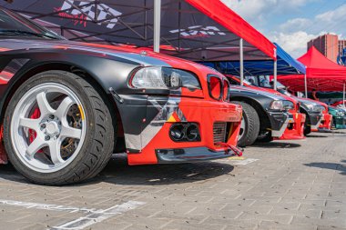 Kırmızı BMW E30 M3, siyah beyaz grafiklerle bir araba fuarında, kırmızı çadırların altında, diğer park halindeki arabaların arasında, Sunny Day, Kyiv, Ukrayna 06-15-2025