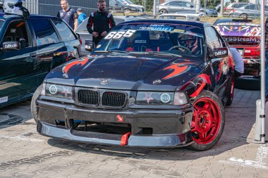 Açık hava araba yarışları, kırmızı, turuncu ve siyah çizgili BMW yarış arabası, 19 numara, sponsor çıkartmaları, geniş arka kanat, düşük duruş, dinamik gölgeler, metalik parıltı, sert Kyiv, Ukrayna 06-15-2025