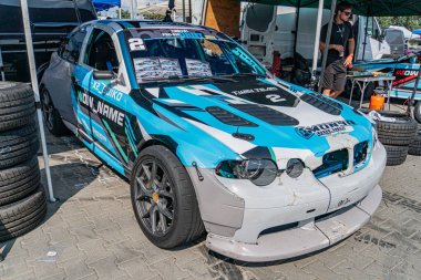 Tuğla kaldırımda sürüklenen mavi BMW, muhtemelen bir garajda ya da pit stop alanında hasarlı ön tampon, yarış lastikleri, sponsorluk logoları, doğal ışıklandırma, gizli stil Kyiv, Ukrayna 06-15-2025