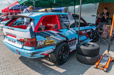 Kyiv Araba Sürükleme Festivali tuğla yolda. Modifiye edilmiş mavi-beyaz kompakt araba, arka spoiler, lastikler, yeşil tepe örtüsü, iki kişi, gün ışığı, canlı renkler, cand Kyiv, Ukrayna 06-15-2025