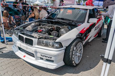 Motorsporlarında pistte beyaz BMW yarış arabası, açık motor kapağı, TUNERZ markası, siyah ve kırmızı yarış üniforması, üstten aydınlatma, uzun gölgeler, izleyiciler, casua Kyiv, Ukrayna 06-15-2025