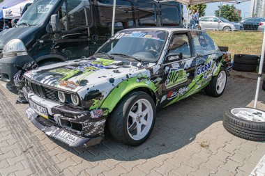 Sürüklenen bir festivalde mavi-yeşil BMW hatchback hasar görmüş, kısmen bir çadır yapısıyla gizlenmiş. Önemli bir ön taraf çarpışma hasarı, buruşuk kaput, parçalanmış headli Kyiv, Ukrayna 06-15-2025