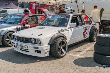 Beyaz BMW yarış arabası Kyiv 'de bir araba festivalinde, ek aerodinamik elementler ve siyah grafik asfalt ayarı, arka planda görünen diğer arabalar gündüz aydınlatması, Kyiv, Ukrayna 06-15-2025