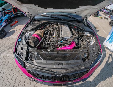 BMW arabanın kapalı kaputu, hortumlar, radyatör fanı gibi motor bileşenleri, ve tuğla döşeli alanda pembe bir ipucu muhtemelen bir garaj ya da Kyiv, Ukrayna 06-15-2025