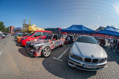 Şehir dışında gündüz saatlerinde gümüş renkli BMW, güneş ışığını yansıtan bir caddeye park etmiş, yakınlarda modifiye edilmiş yarış arabaları ve park halindeki araçlar Kyiv, Ukrayna 06-15-2025