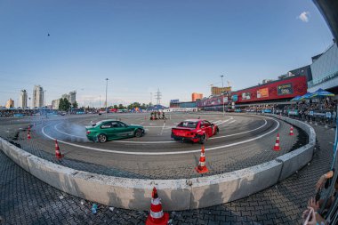 Beyaz çizgilerle işaretlenmiş beton bir yolda sürüklenen kırmızı ve beyaz arabalar tribünlerdeki izleyicilerle çevrili. Lastiklerden duman görünüyor. Şehir manzarası arka planı Kyiv, Ukrayna 06-15-2025