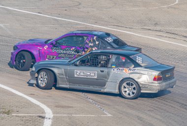 Kyiv Araba Sürükleme Festivali sahnesi yarış pistinde iki modifiye BMW arabası, biri mor diğeri gri, beyaz işaretli Asphalt pisti Kyiv, Ukrayna 06-15-2025