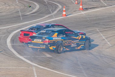 Mavi ve sarı BMW, kırmızı ve siyah takip, araba sürüklenme festivali, asfalt yarış pisti, sola dönüş, toz bulutu, turuncu trafik konisi, gün ışığı, dinamik sahne Kyiv, Ukrayna 06-15-2025