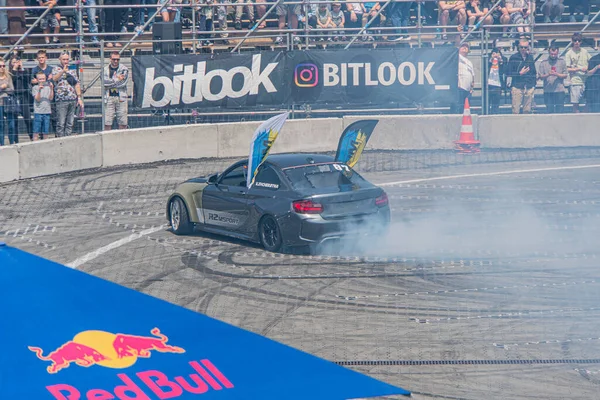 BMW arabası, Kyiv Araba Sürükleme Festivali sırasında, izleyiciler ve markalı pankartlarla çevrili bir yarış pistinde hareket halinde. Araba duman saçıyor, agresif bir duruşu var.