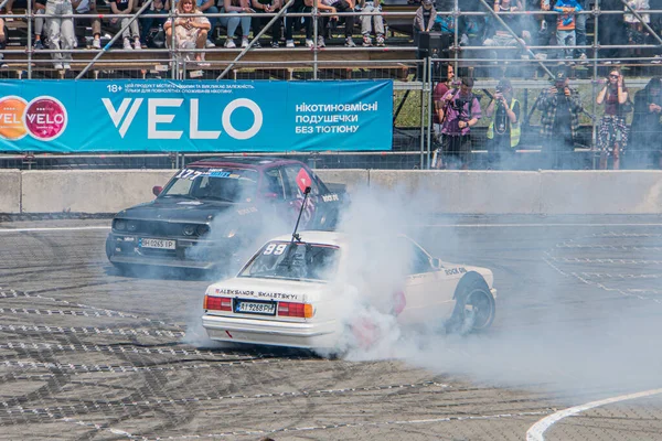 BMW arabası Kyiv Araba Sürükleme Festivali 'nde sürükleniyor. İki yüksek performanslı araba bir pistte ilerliyor, lastik dumanı üretiyor. Yan ve arka camlarda çıkartmalar var.