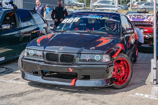 Açık hava araba yarışları, kırmızı, turuncu ve siyah çizgili BMW yarış arabası, 19 numara, sponsor çıkartmaları, geniş arka kanat, düşük duruş, dinamik gölgeler, metalik parıltı, sert Kyiv, Ukrayna 06-15-2025