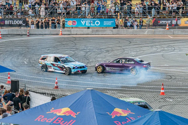 Kyiv Araba Sürüklenme Festivali 'nde süratli sürüklenme yarışı, iki BMW mavi-beyaz, pist üzerinde mor yarış, seyirciler batan güneş gibi çitten izliyor. Kyiv, Ukrayna 06-15-2025