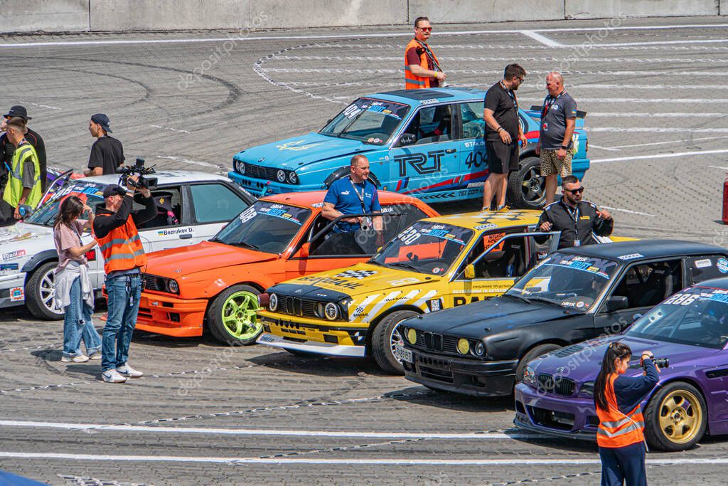 Racers gathered at a bustling concrete race track, vibrant racing cars arranged in rows, pit stop or pre-race preparation area, bright daylight Kyiv, Ukraine 06-15-2025