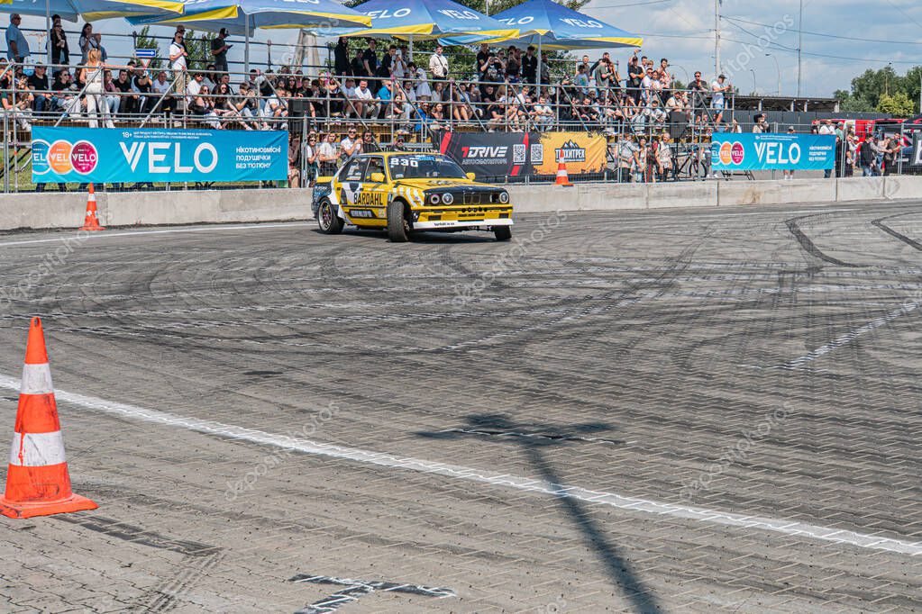 Bright yellow BMW leading at Kyiv Car Drifting Festival on dark asphalt track Scattered safety measures, blurred crowd, daylight, candid style Kyiv, Ukraine 06-15-2025
