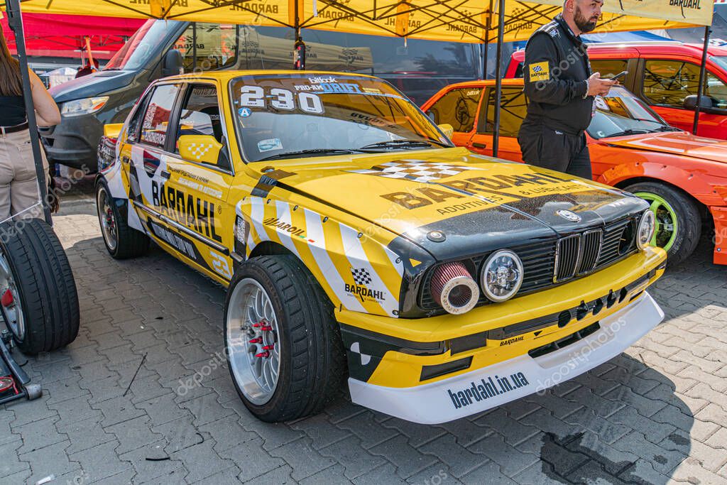 Race-modified BMW E30 yellow, black at car show under yellow tent Number 230, Budweiser sponsor Natural daylight, multiple people in background Kyiv, Ukraine 06-15-2025
