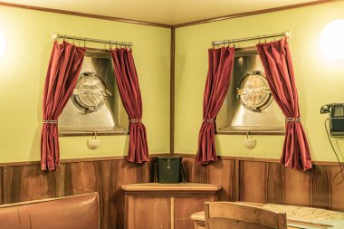 Bir savaş gemisi kabininin içi, ahşap paneller, kravatlı kırmızı perdeler, pencereli ya da yuvarlak pencere, okunamayan bir levha, iki ahşap sandalye, yapay ışıklandırma, faydacı stili