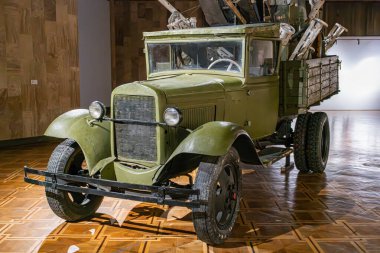 Kaputu açık ve iyi bakımlı yeşil askeri bir kamyonun kapalı müze vitrininde büyük farları, belirgin ızgarası, ağır çamurlukları, yedek lastiği var.