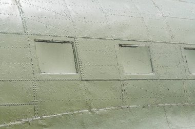 Üç dikey pencereli koyu yeşil bir uçak gövdesinin yakın görüntüsü aşınma ve aşınma belirtileri gösteriyor. Görüntü yer seviyesinden alınıyor ve uçağın büyüklüğünü vurguluyor.