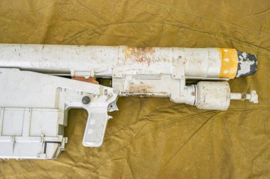 Bir ucunda kırmızı ve sarı kesitler olan beyaz, silindirik bir nesne, muhtemelen bir oyuncak ya da model silah, kumaş arkaplanının üzerine yerleştirilmiş, metin ya da insan figürü yok