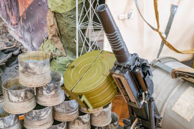 Ağır bir askeri top mermisi parçasının, istiflenmiş cephane kutularının üzerinde durduğu kapalı bir mekan, geçici bir barınak ya da depolama alanı, sessiz tonlu, gölgeli, dünyevi renk paleti ve...