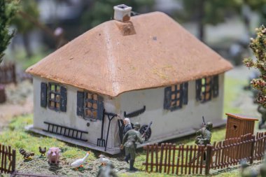 Sazlı bir kır evi, domuz, horoz ya da tavuk figürleri, yeşil yapraklar, toprak yol, ahşap çit, doğal renkler, hatta ışıklandırma olan minyatür model köy sahnesi.