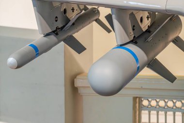 Gümüş renkli, mavi çizgili, beyaz şapkalı bir füzenin yakından görüntüsü, bir hava aracının altına monte edilmiş, kapalı alanda, yapay ışıklandırma.