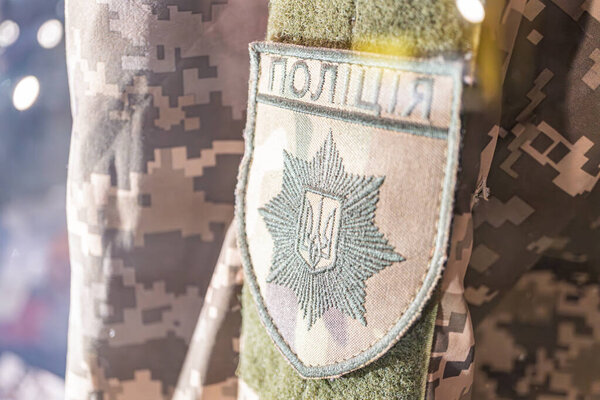 Рука видна с камуфляжным рукавом, спортивные два патча Один патч содержит текст жирными буквами, окруженный венком, и другое слово, начинающееся с, как на военном, так и на офисном